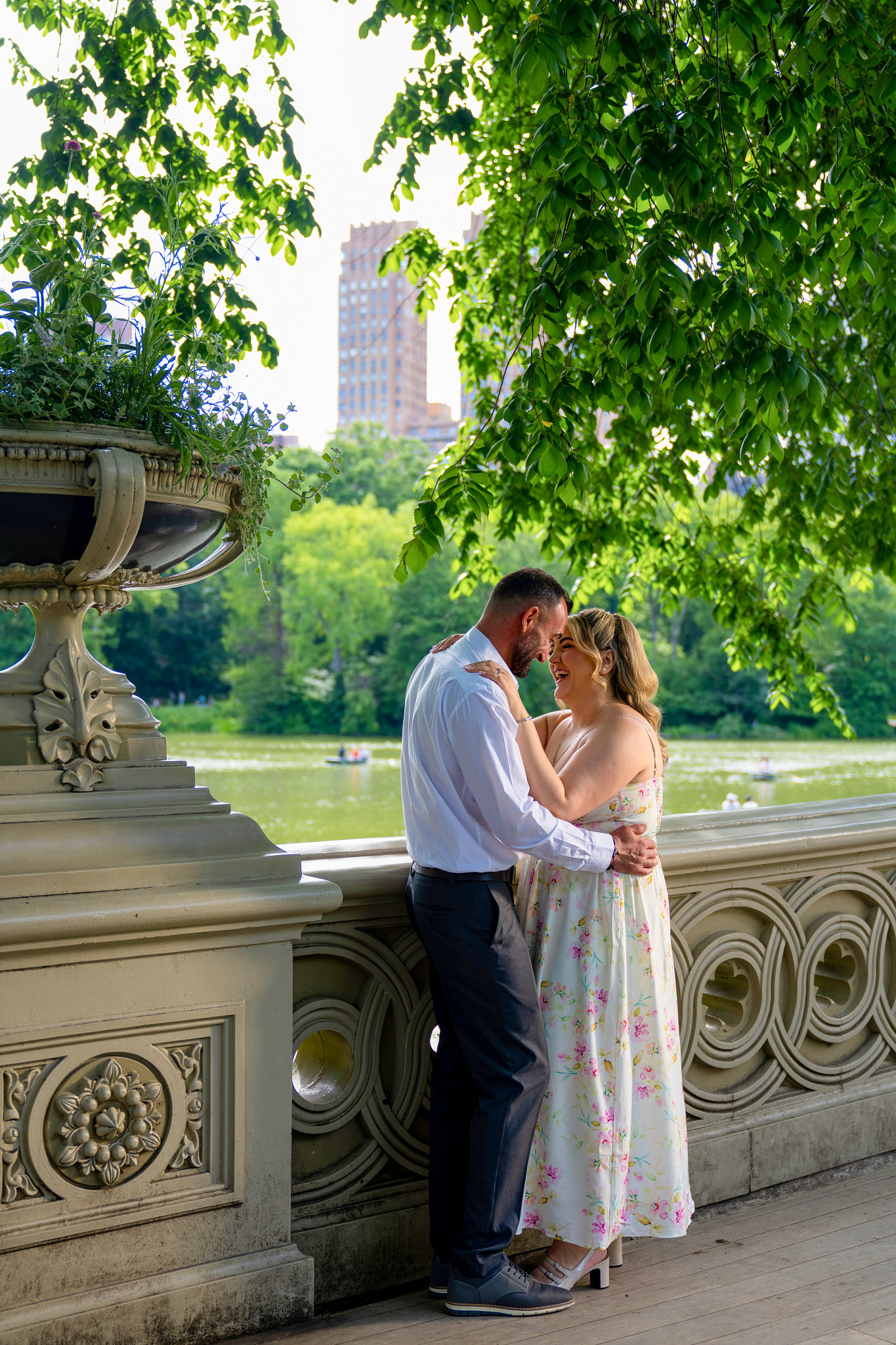 Couple embracing and laughing on a leafy lakeside bridge in a city park.